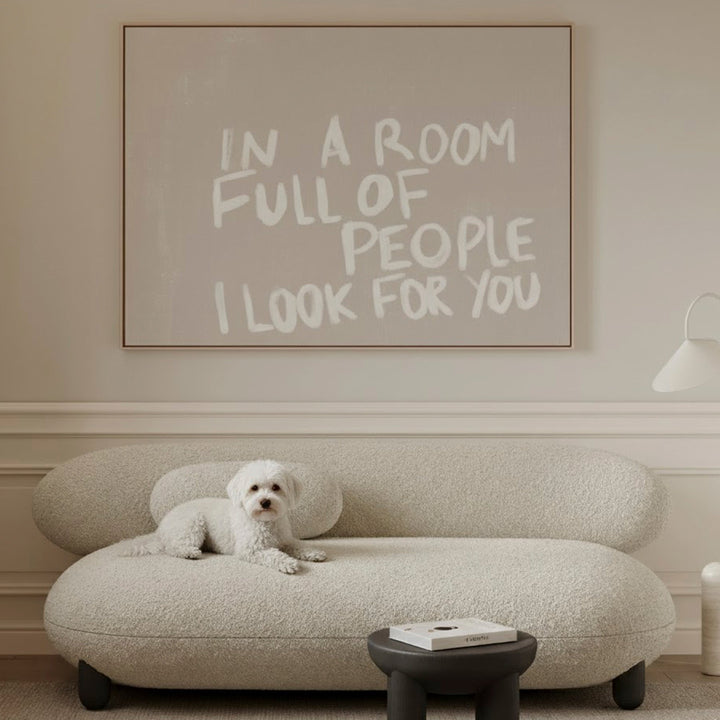 White dog on a modern beige sofa with a framed quote in a living room, there is a large framed canvas with a beige background and white handwritten style text which reads "in a room full of people I look for you"