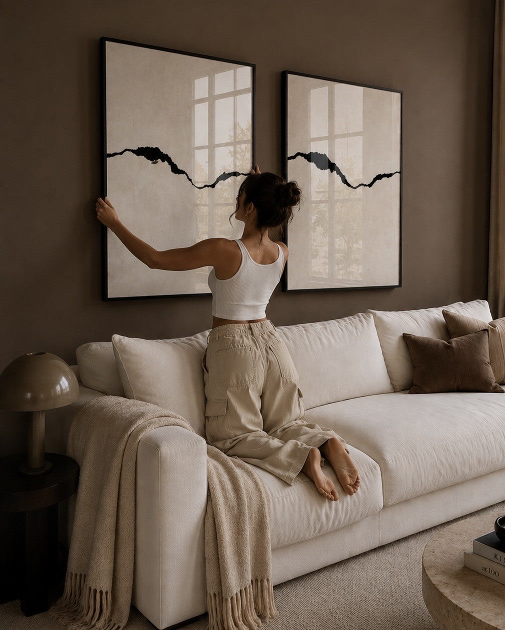 A woman kneels on a cream-colored sofa in a modern living room, adjusting a large black and white abstract framed print on a brown wall. The room is styled with neutral tones, including a cream sofa, a beige throw blanket, and matching pillows.