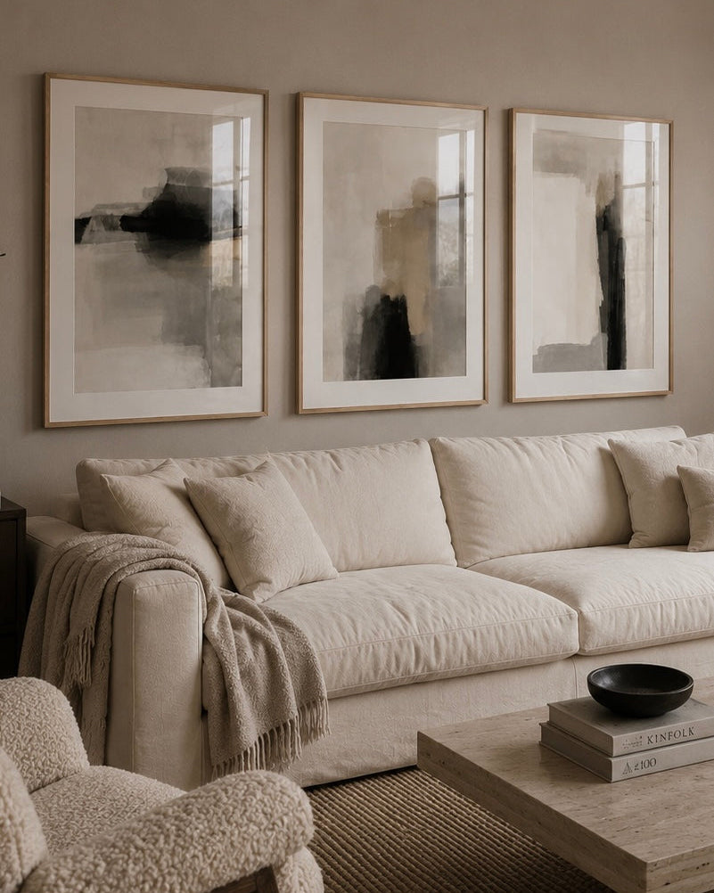 A styled living room featuring three large framed abstract black and gray wall art prints hanging above a cream-colored fabric sofa. The room has a warm neutral aesthetic with a matching textured armchair in the foreground, a light wood coffee table, and a woven rug.
