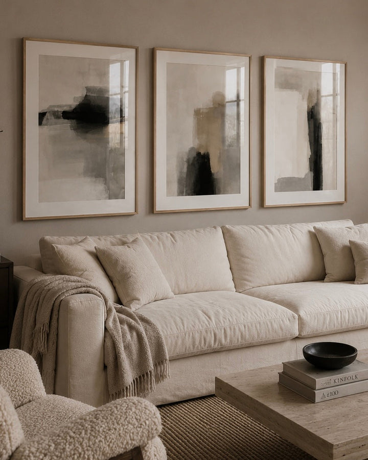 A styled living room featuring three large framed abstract black and gray wall art prints hanging above a cream-colored fabric sofa. The room has a warm neutral aesthetic with a matching textured armchair in the foreground, a light wood coffee table, and a woven rug.