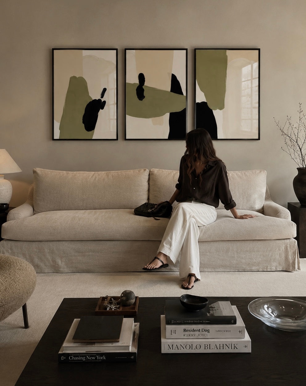 A woman sits on a beige sofa in a styled living room, with three framed abstract wall art prints above the couch. The artwork features soft sage green, beige, and black shapes on a light background.