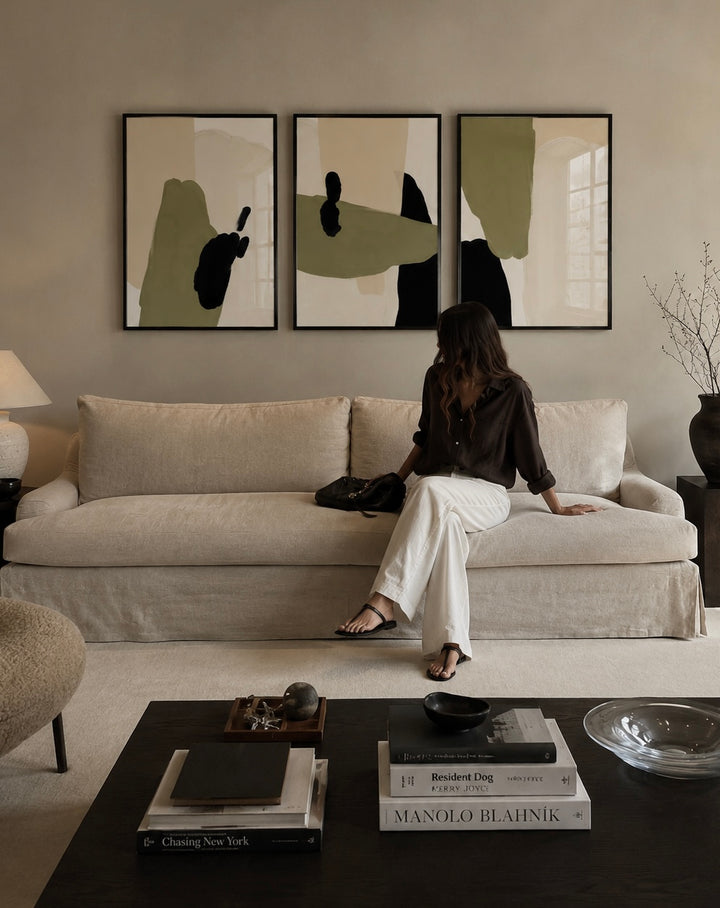 A woman sits on a beige sofa in a styled living room, with three framed abstract wall art prints above the couch. The artwork features soft sage green, beige, and black shapes on a light background.