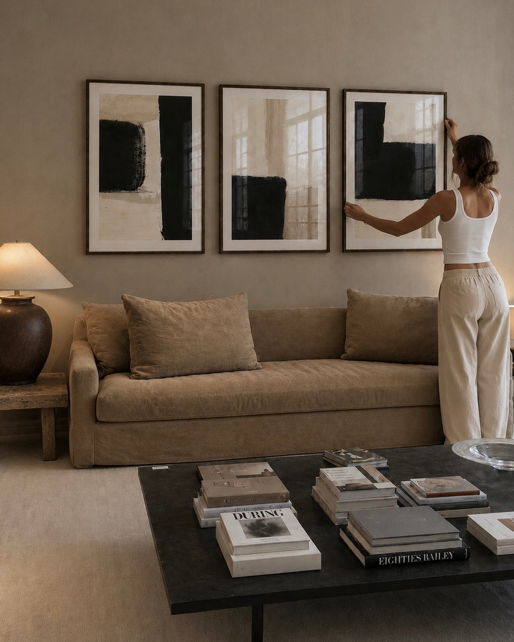 A woman in a white tank top and beige pants stands beside a beige sofa in a modern living room, touching a large black-and-white framed abstract print hanging on the wall. A matching sofa, lamp, and coffee table are visible in the room.