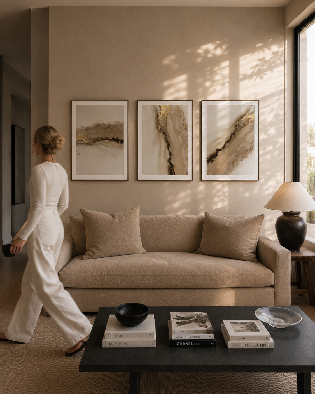 A woman in a white outfit walks past a beige sofa in a modern living room. Three framed abstract artworks hang on the wall above the sofa, and a black coffee table with books and decor sits in front.