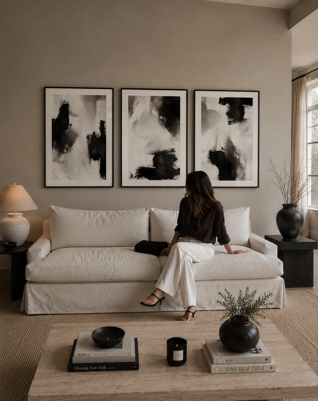 A woman sits on a cream slipcovered sofa in a styled living room, facing three large framed abstract black-and-white wall art prints. The room has warm taupe walls, a light wood coffee table, and dark ceramic vases.
