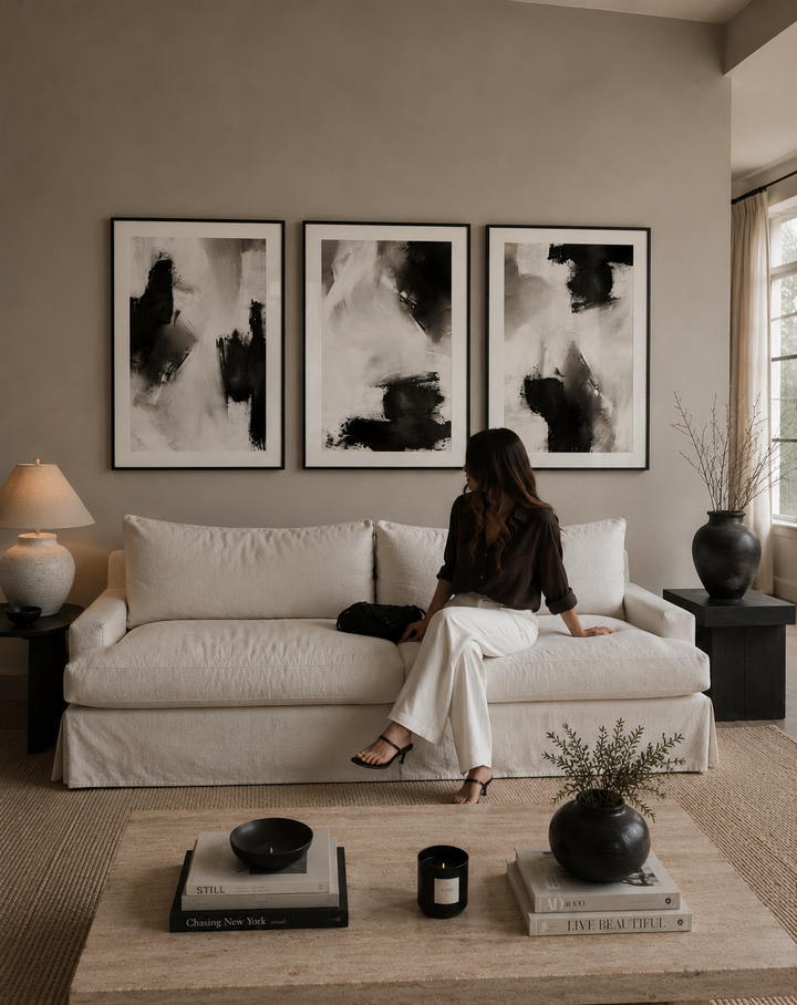 A woman sits on a cream slipcovered sofa in a styled living room, facing three large framed abstract black-and-white wall art prints. The room has warm taupe walls, a light wood coffee table, and dark ceramic vases.