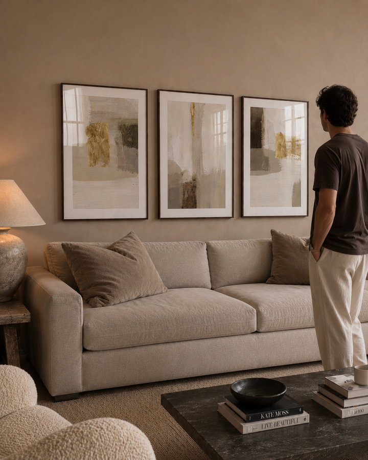 A man stands in a living room looking at a set of three framed abstract wall art prints hanging above a beige sofa. The room has warm neutral tones, a textured lamp, and a dark wood coffee table.