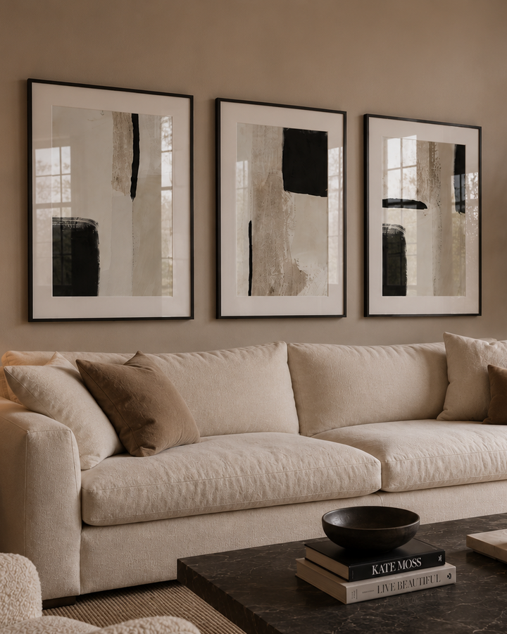 A beige upholstered sofa in a styled living room, decorated with three framed abstract black-and-white wall art prints, a table lamp, a dark coffee table, and a potted plant.