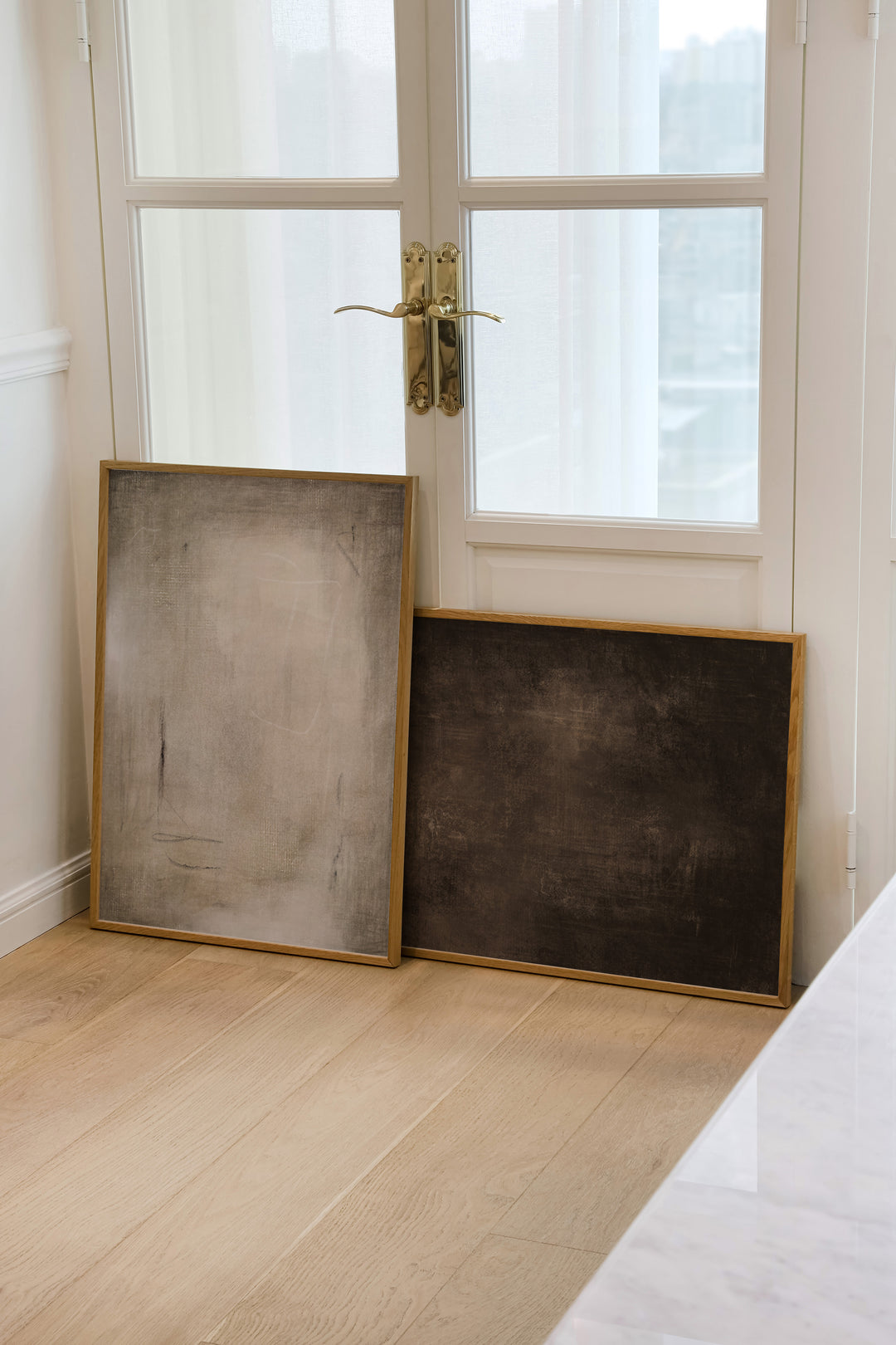 Two framed artwork prints with a neutral colour palette of brown and beige on a wooden floor with a glass door in the background.
