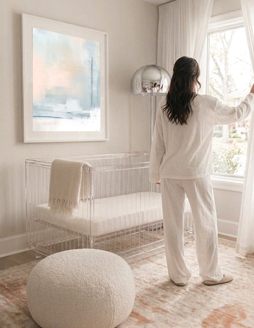 Woman in a white outfit standing by a window in a bright room with a crib and framed artwork.