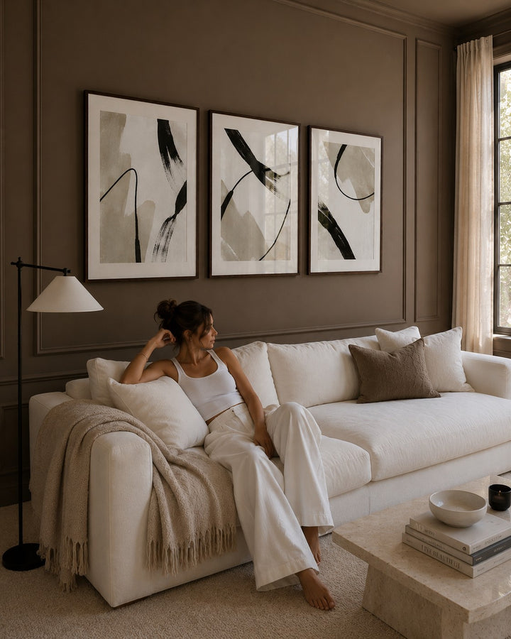 A woman sits on a white sofa in a styled living room, wearing a white tank top and white wide-leg pants. The room features a warm brown wall with three framed abstract black, white, and gray prints, a beige throw blanket, and a stone coffee table.